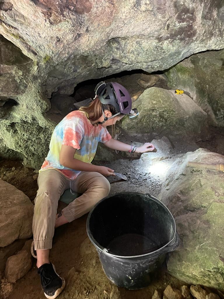 Jasmine is sitting down excavating in a cave setting.