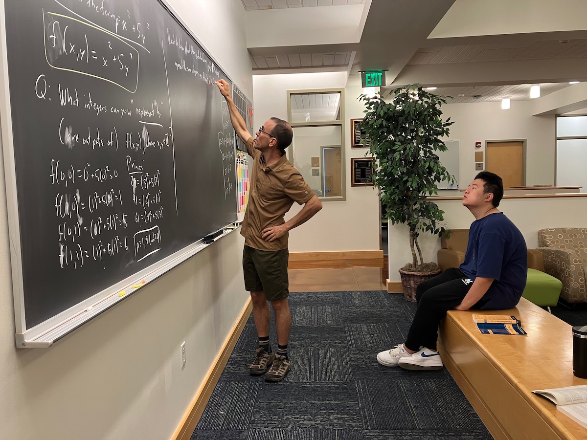 A man writing algebra functions on a whiteboard while a college student observes