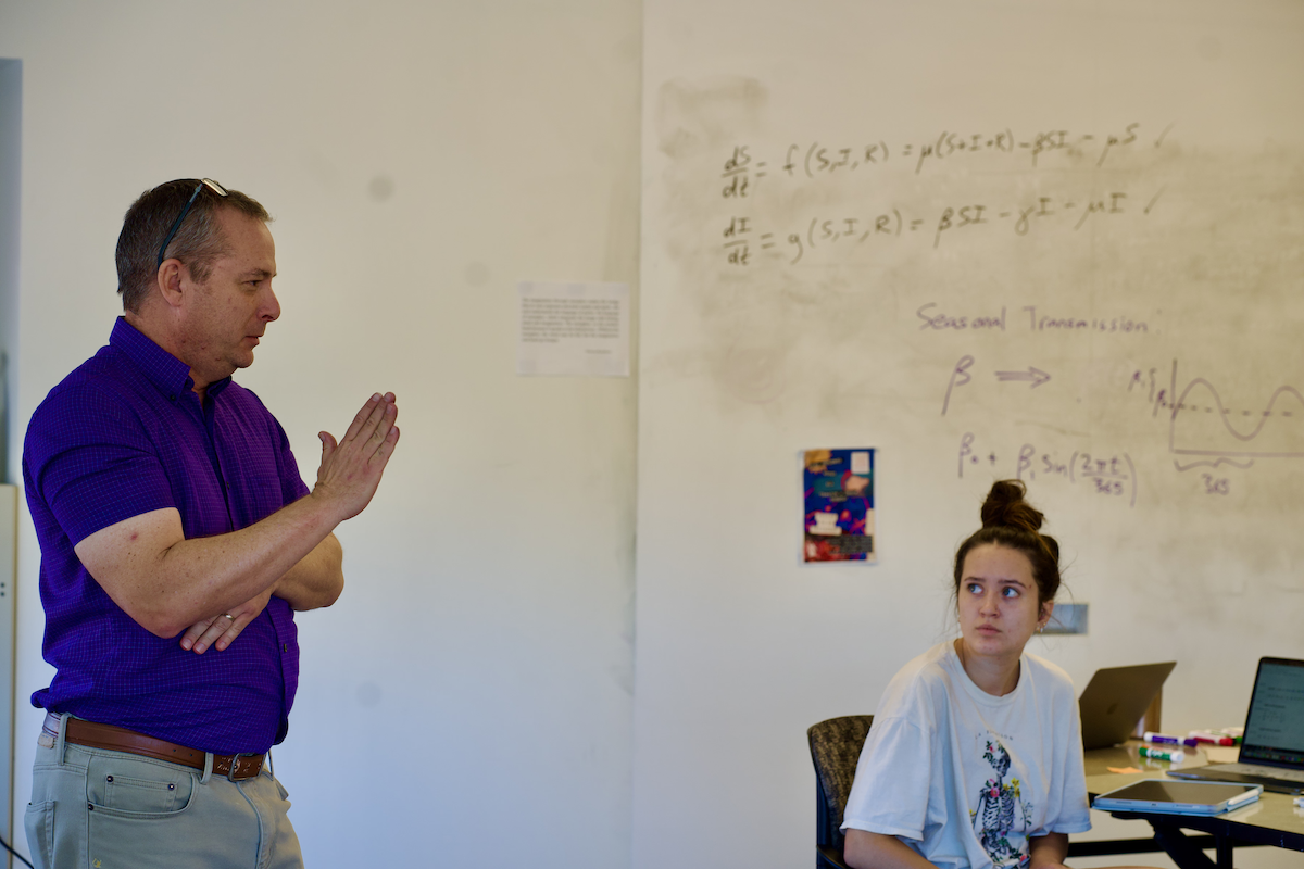 A male professor in a blue dress shirt explaining a concept of a class