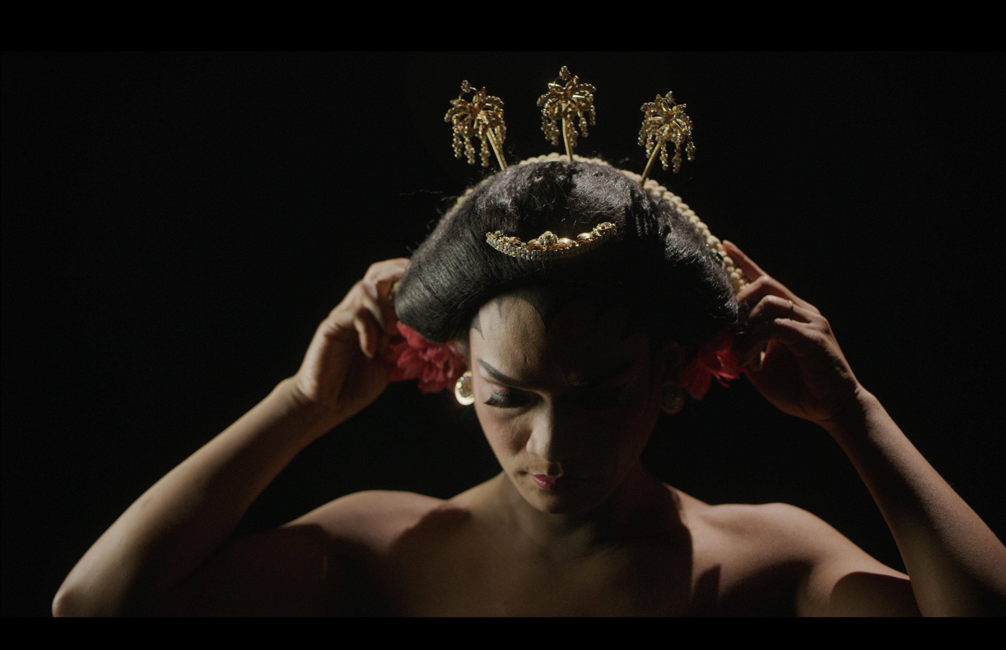 Rianto adjusting an ornate traditional headdress and red floral adornments against a dark background.