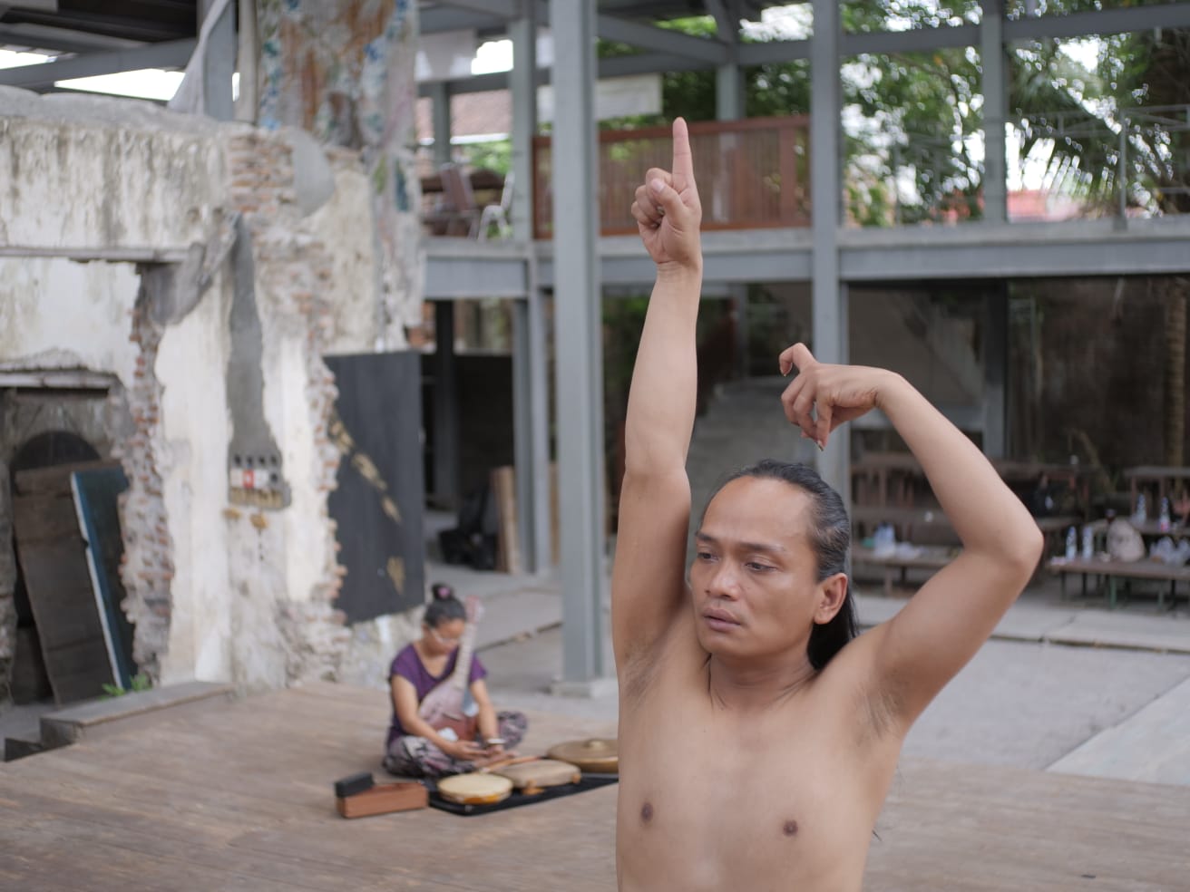 Rianto performing a dance gesture with raised arms in an open-air performance space, with a musician seated in the background.