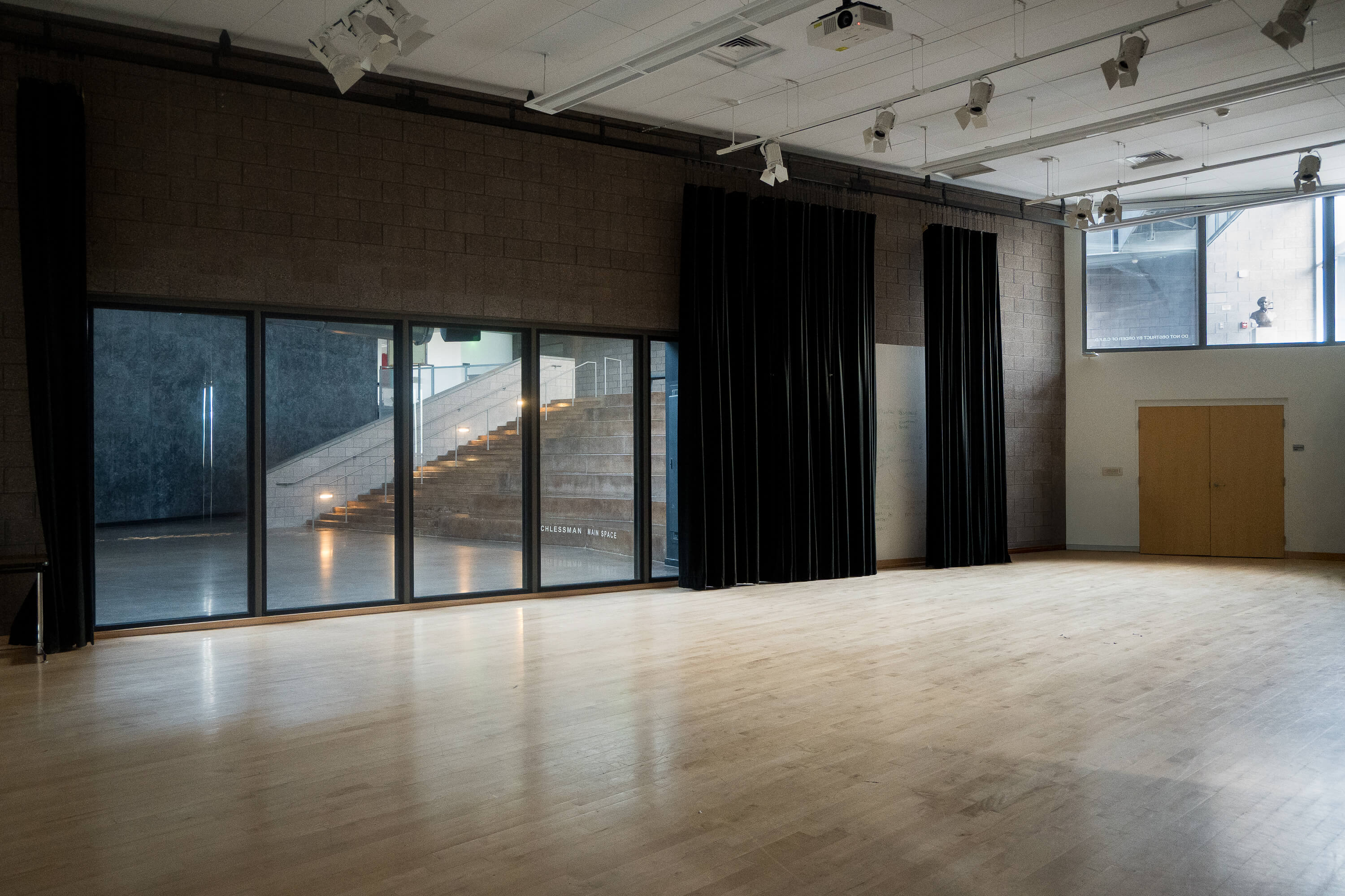 Interior of the Flex Room, featuring an open wood floor, black curtains, overhead lighting, and large windows overlooking the adjacent performance space.