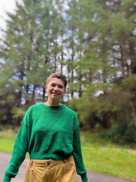 Abi smiling outdoors in a green sweater and tan pants, standing in front of a wooded background.