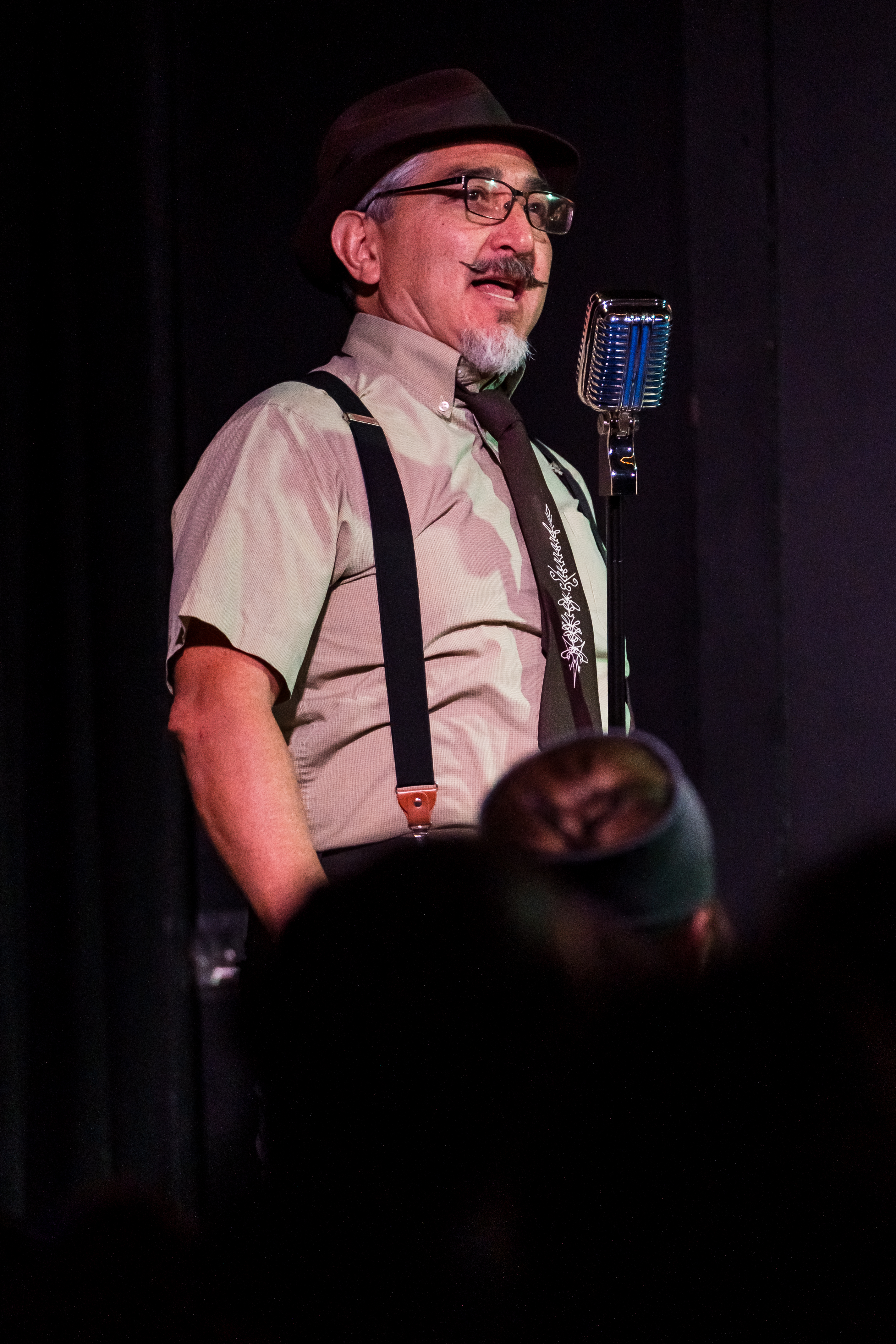 Adrian Holguin performs onstage, wearing a black fedora, glasses, suspenders, and a tie, while speaking into a vintage-style microphone.