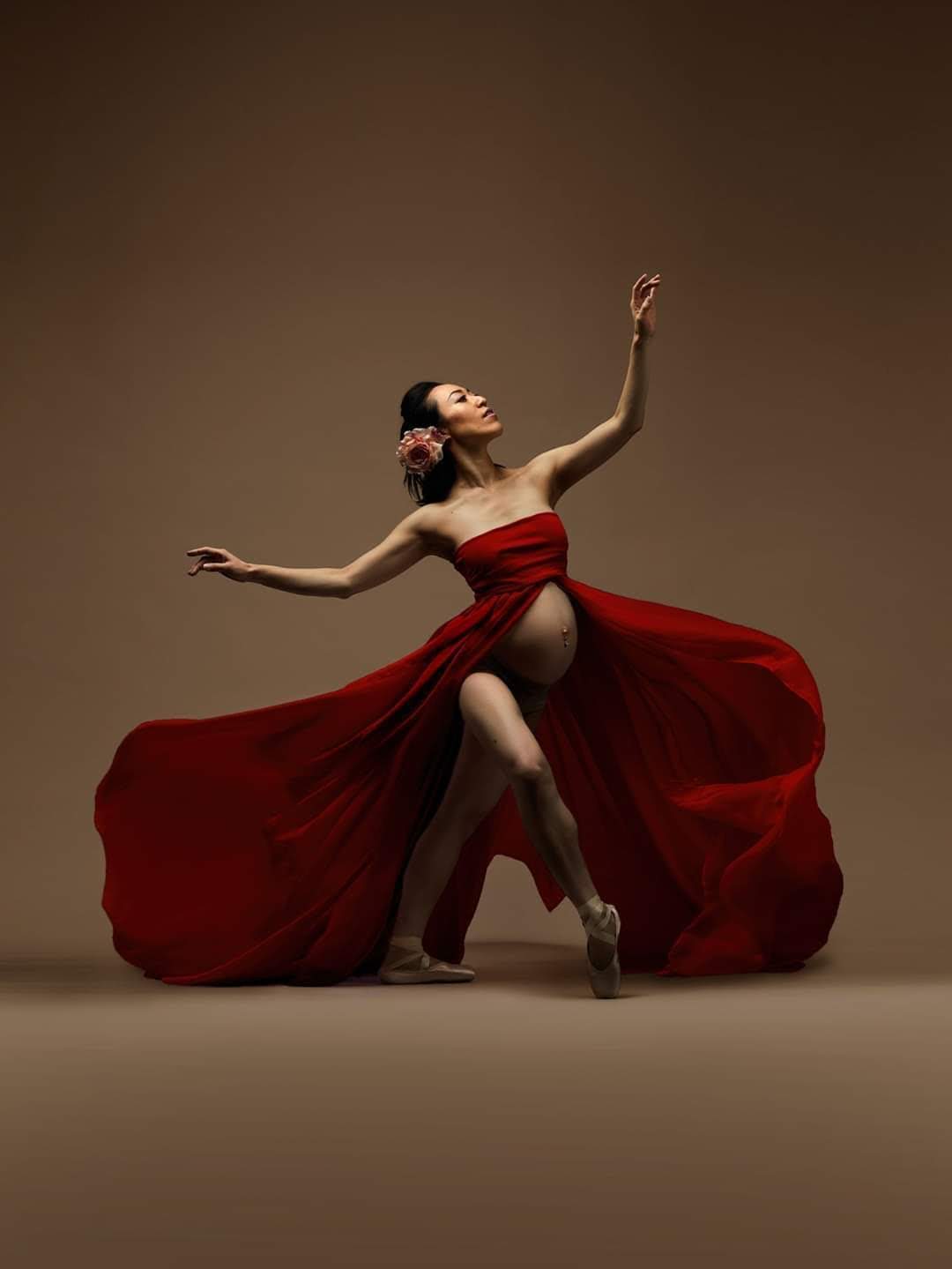 Aoi Koenig posing en pointe in a flowing red dress with a flower in her hair, extending her arms in a graceful dance pose.