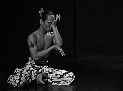 Rianto seated in a dance pose, wearing patterned fabric against a dark background.