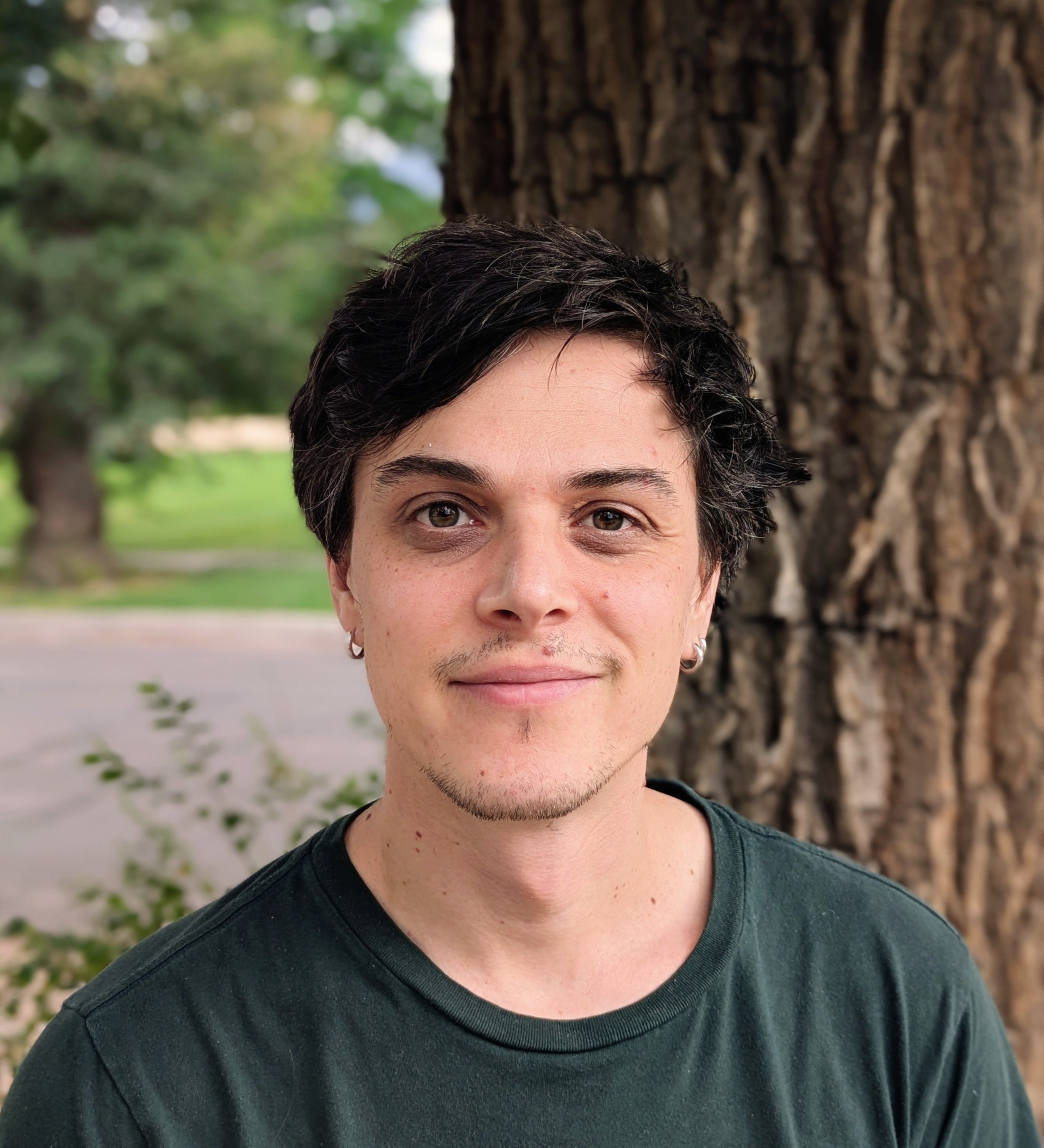 Ruslan Khusein smiling at the camera in a dark green shirt, standing outdoors in front of a large tree.