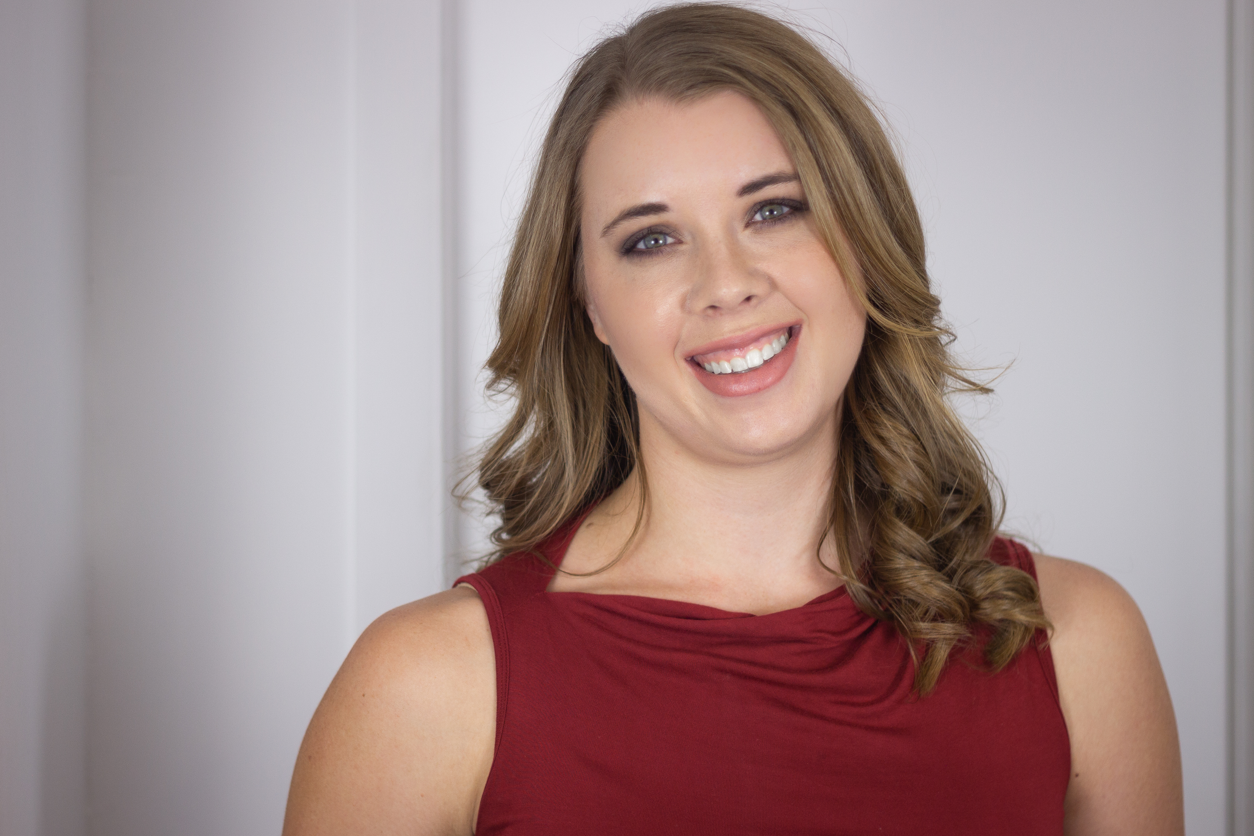 Headshot of a smiling Samantha Egle with shoulder-length wavy brown hair, wearing a sleeveless red top against a light gray background.