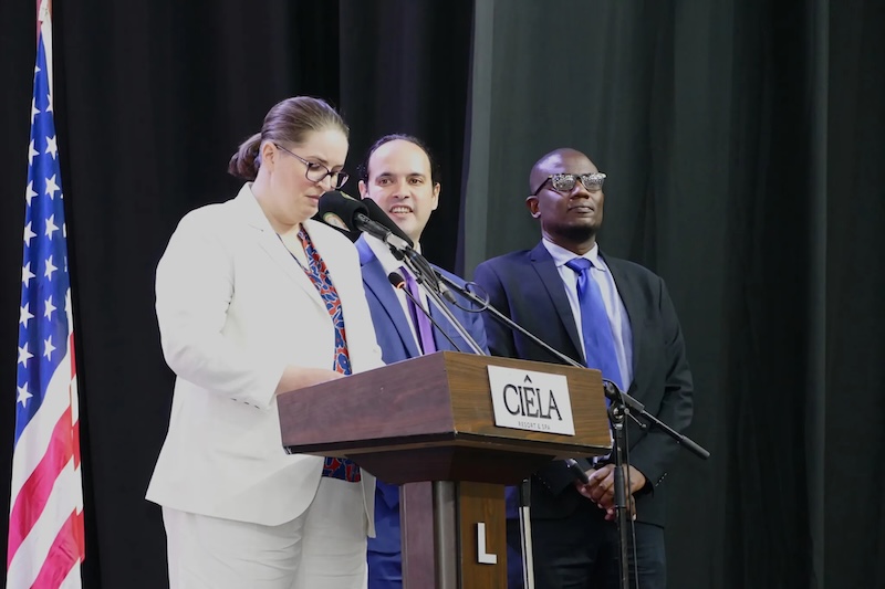 Collinson presenting at a conference in Zambia. She stands behind a podium and microphone. There's an American flag and two men on stage with her in view.