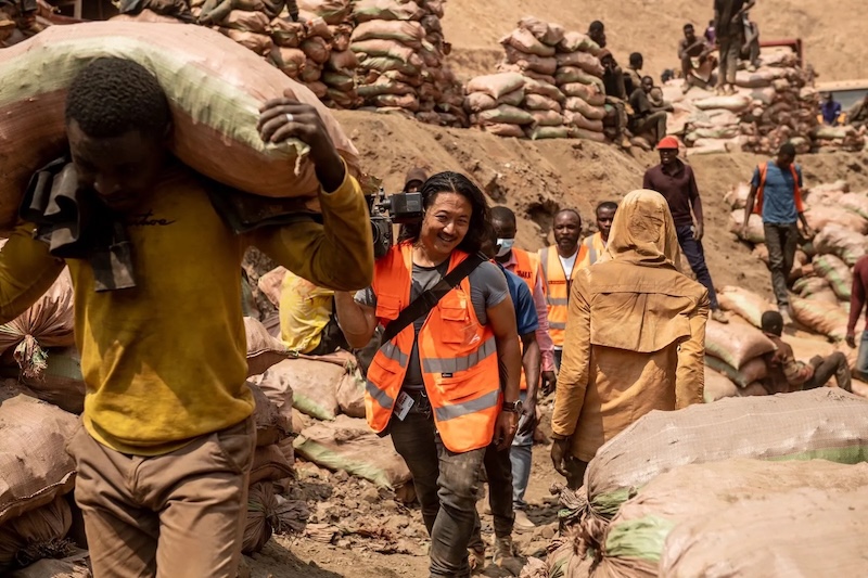 Mike Shum wearing bright orange visibility vest and carrying a video camera walking in line with cobalt miners in the Shabara artisanal mine near Kolwezi in the Democratic Republic of Congo