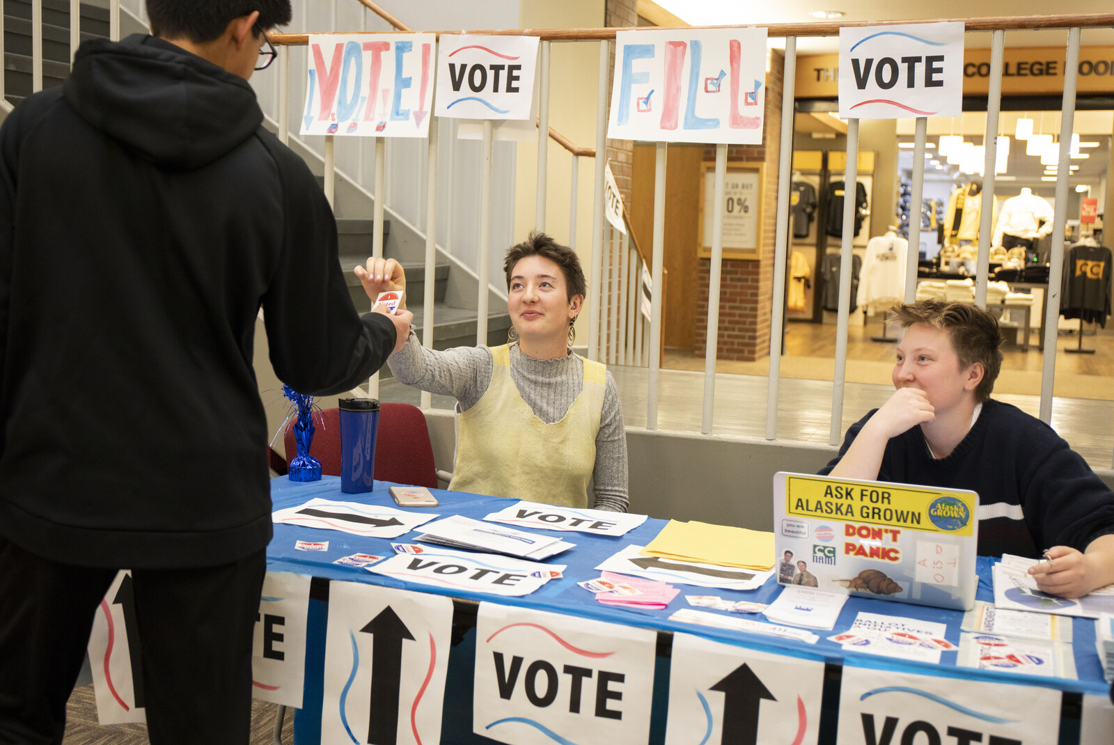 Two student volunteers sit behind a voting desk. One hands an "I voted" sticker to a student at the desk.