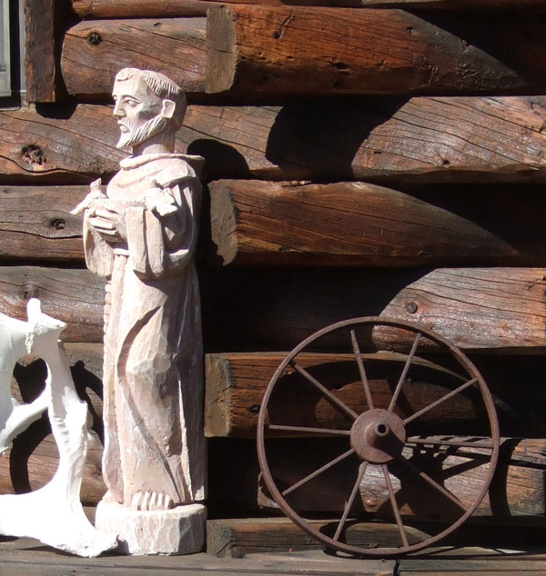 St. Francis statue in front of some wooden logs and next to a rusted wheel