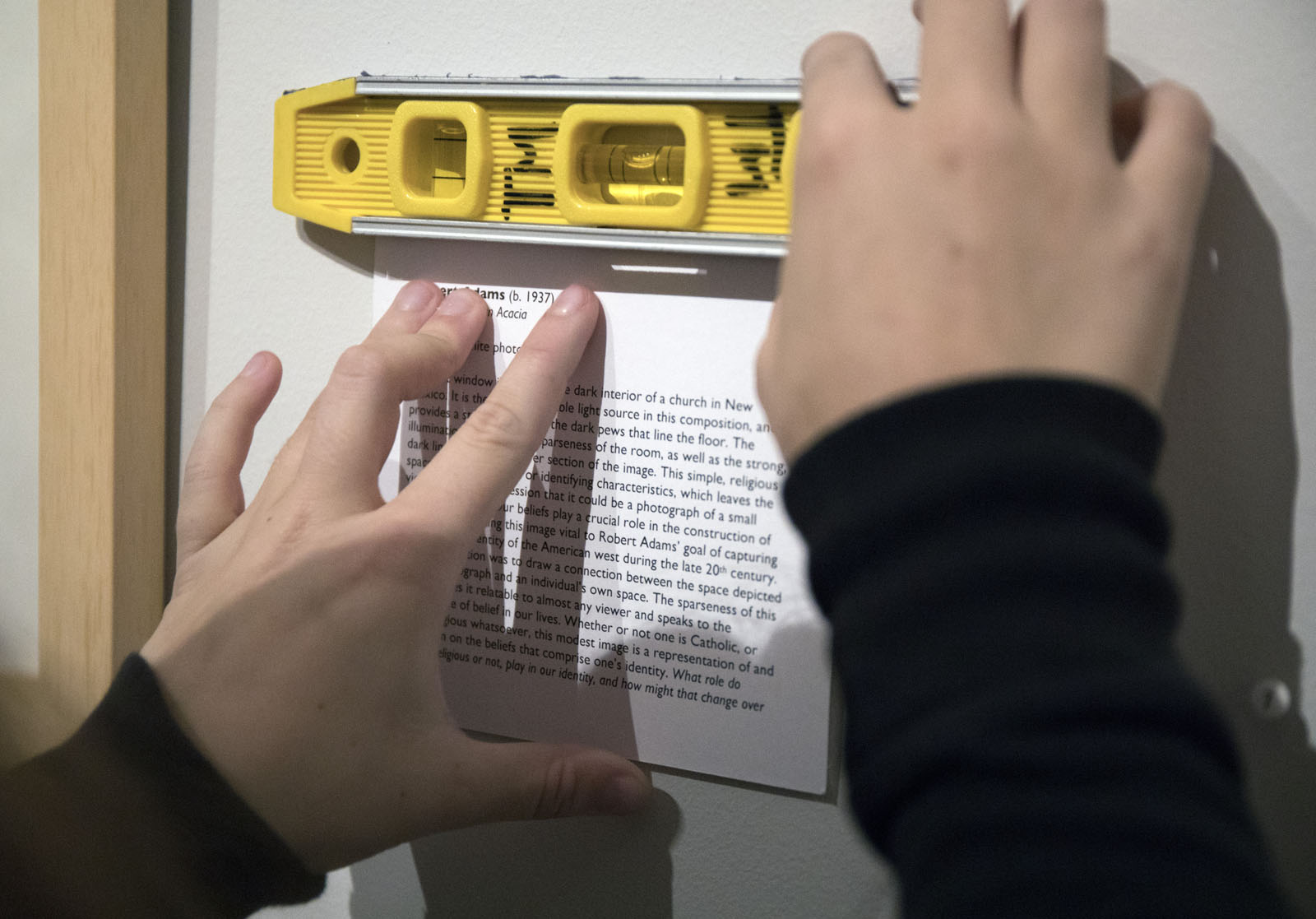 An FYE student uses a level to make sure the description plate that she wrote was level and even on the wall in the exhibit room at the Fine Arts Center.
