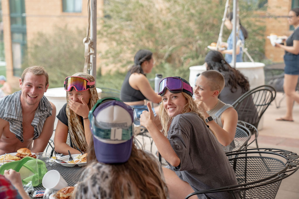 Students smiling and eating at a table with a line of other students in the background <span class="cc-gallery-credit">[Gray Filter Photography]</span>