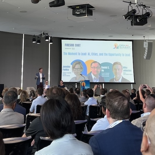 A speaker presents at the front of an auditorium at a conference