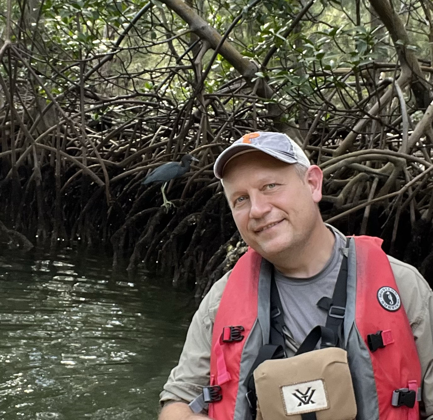 Professor Miero Kimmel wearing a life vest in a river setting. Behind him, a blue bird perches on a branch.